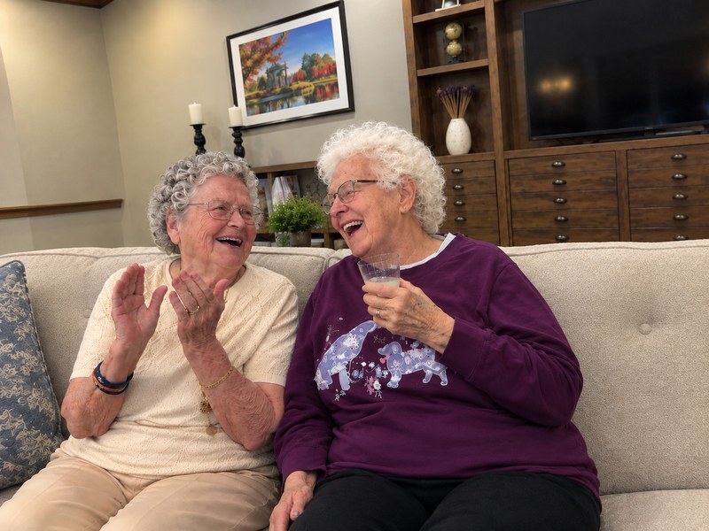 Two assisted living residents laughing together on a sofa in a comfortable common area.