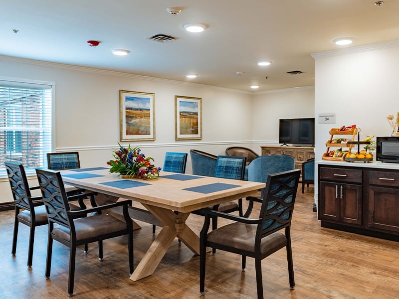 Enjoy this accessible common area at Cedarhurst of Tesson Heights, which has a dining table and comfrtable seating, a TV, a microwave, and a coffee machine