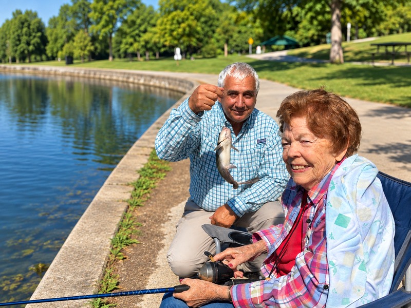 Cedarhurst of Tesson Heights senior living resident fishing by a lake as a companion shows off the catch of the day
