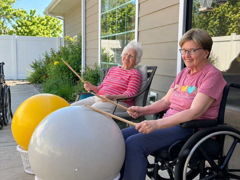 Assisted living residents drumming on exercise balls during an outdoor drum fitness class.