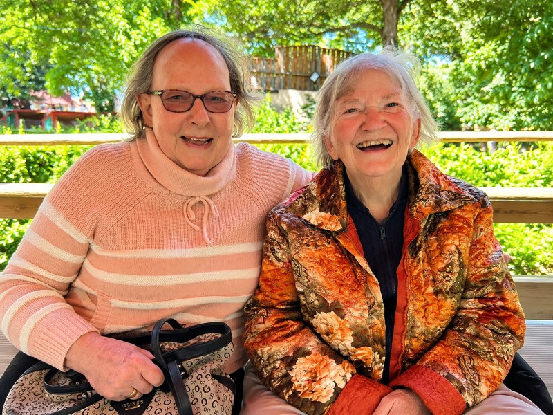 wo senior living residents laughing together on a bench surrounded by greenery.