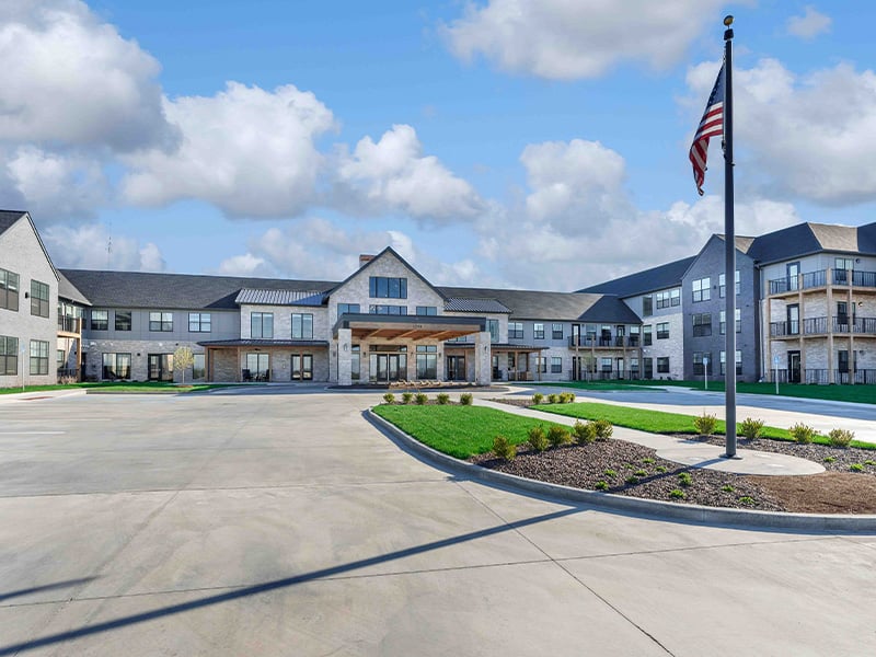 An inviting view of the Cedarhurst of Wentzville senior living community from the outside, complete with the main building, a flag, and the driveway