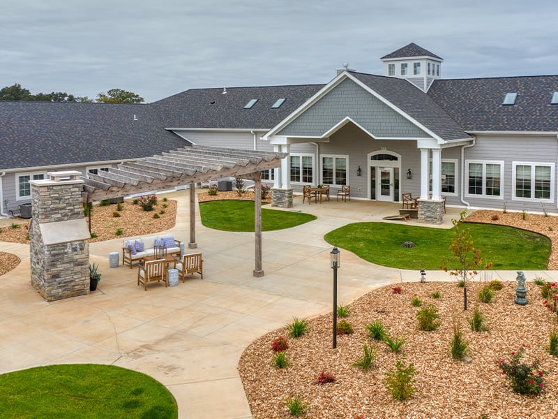An inviting outdoor area at Cedarhurst of West Plains, showing lush green spaces, paved paths, comfortable seating, a pergola, and a covered patio.