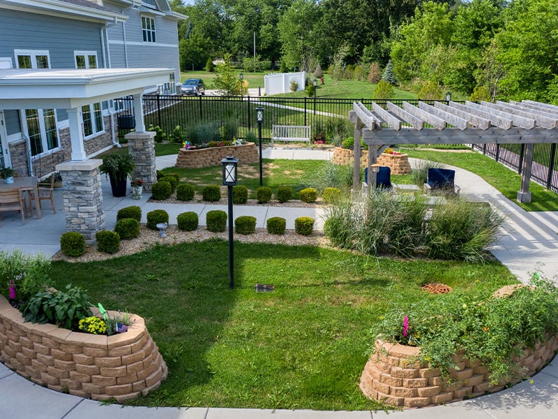 Outdoor green space at Cedarhurst of Yorkville with paved walking paths, a wooden pergola, and raised garden beds