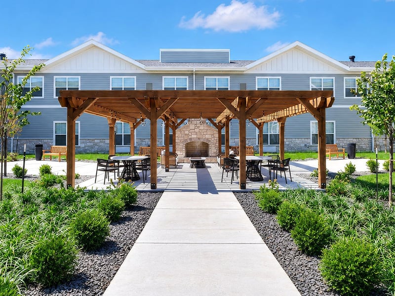 An inviting outdoor area at Cedarhurst of Shawnee, showing lush green spaces, paved paths, comfortable seating, and a pergola