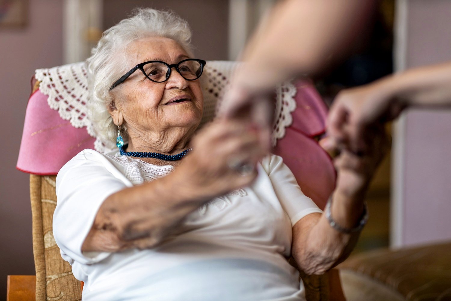 A seated senior woman looking up at a caregiver who is holding her hands