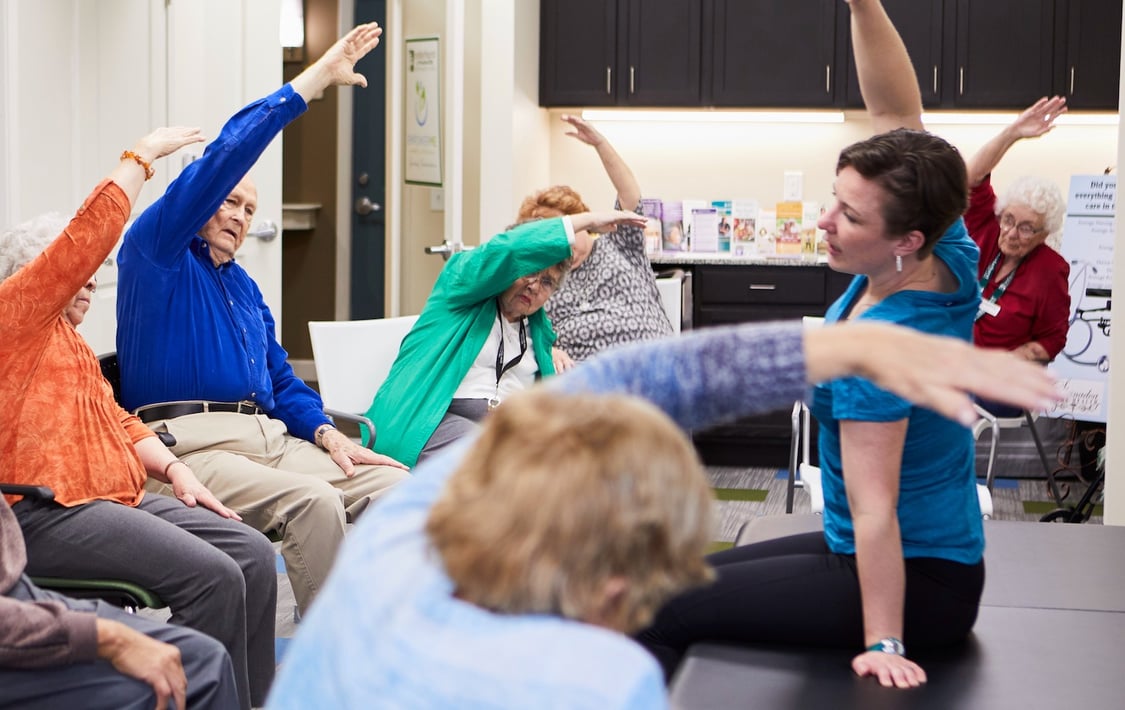 A group of six seniors participating in a seated exercise class led by a staff trainer