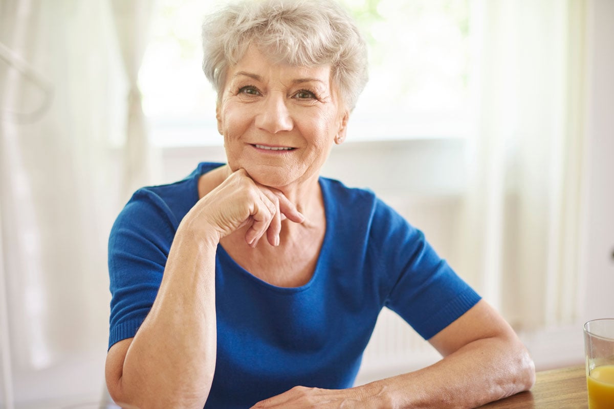 Elderly woman with short gray hair wearing a blue blouse, sitting indoors by a softly lit window, smiling warmly with a hand resting on her chin