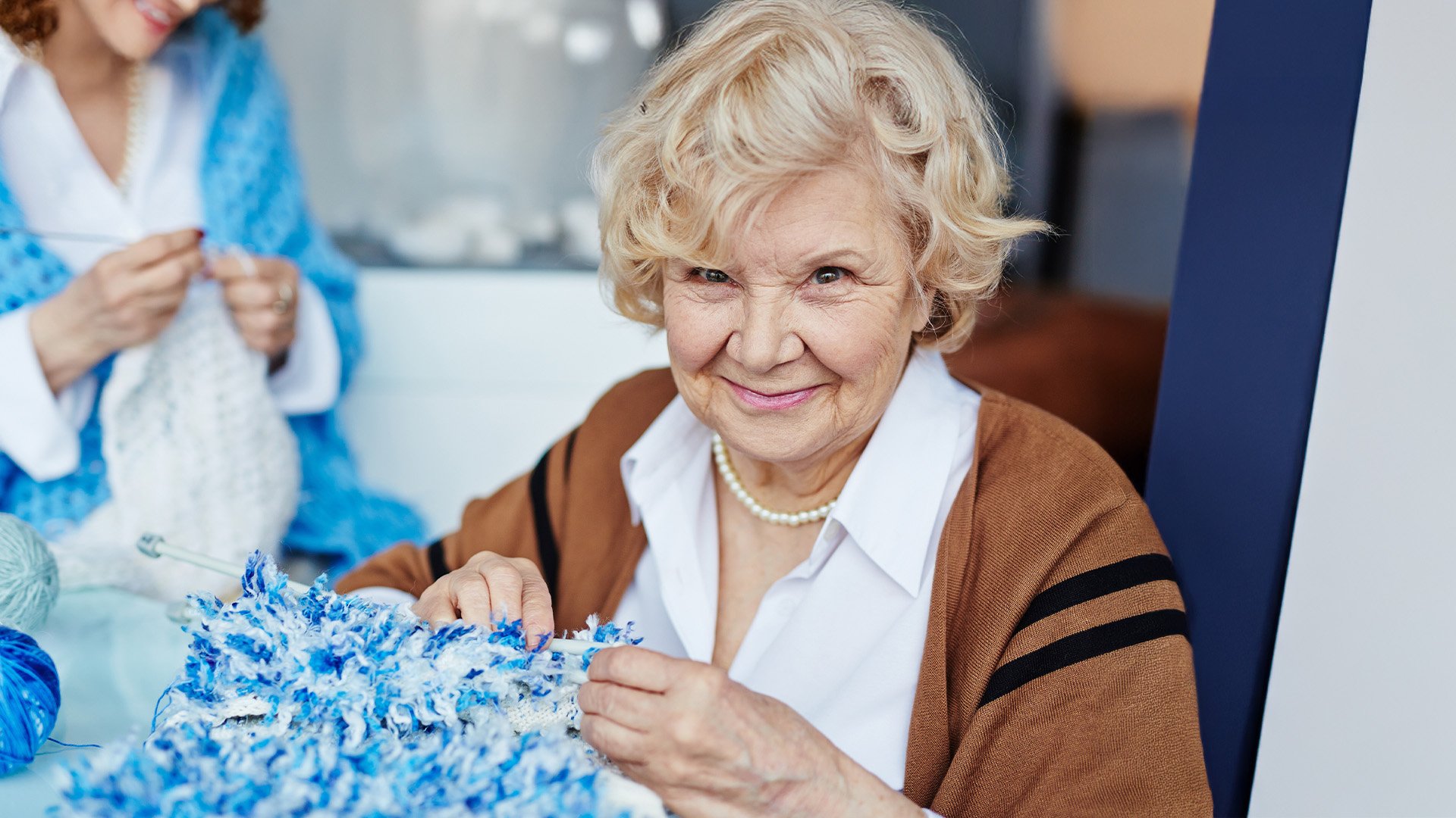 Portrait of senior living resident looking at camera with deep black eyes while holding unfinished knitted scarf and needles in hands