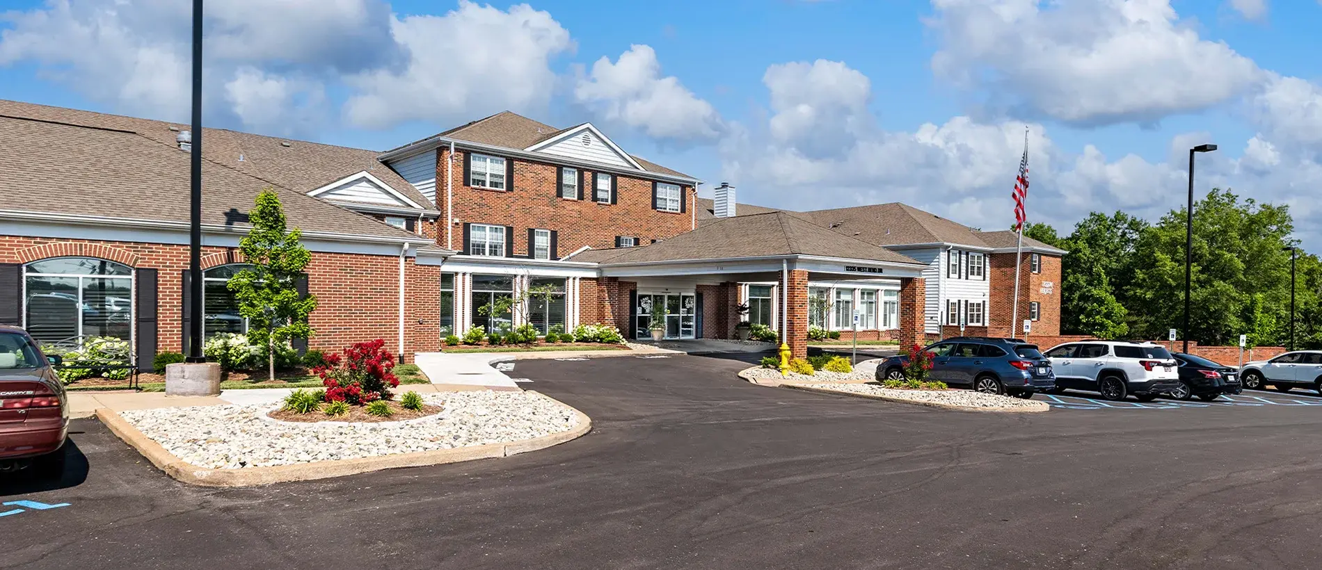 A brick building with multiple windows and a sloped roof, surrounded by a parking area with cars