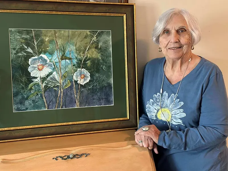 Elderly woman smiling beside a framed floral painting with white flowers and green foliage