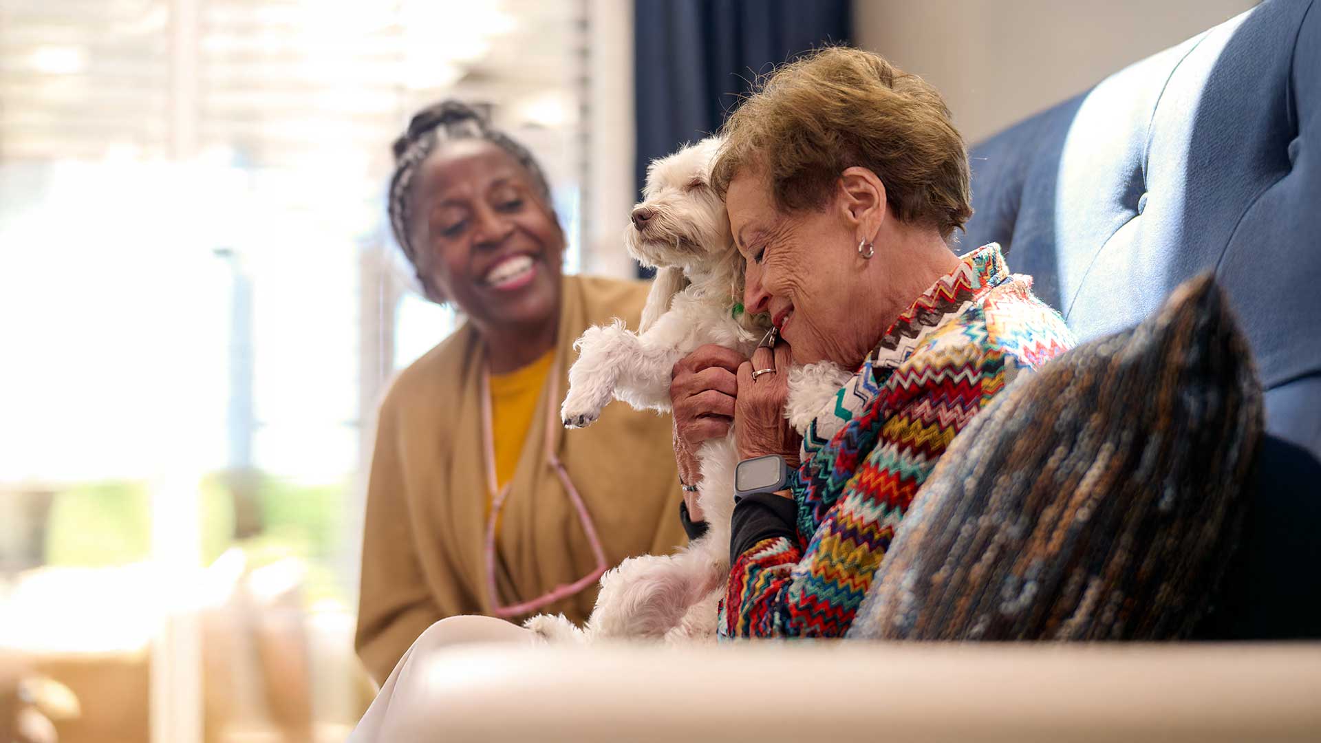 Senior woman sitting on a couch hugging a small white dog while another senior woman smiles in the background