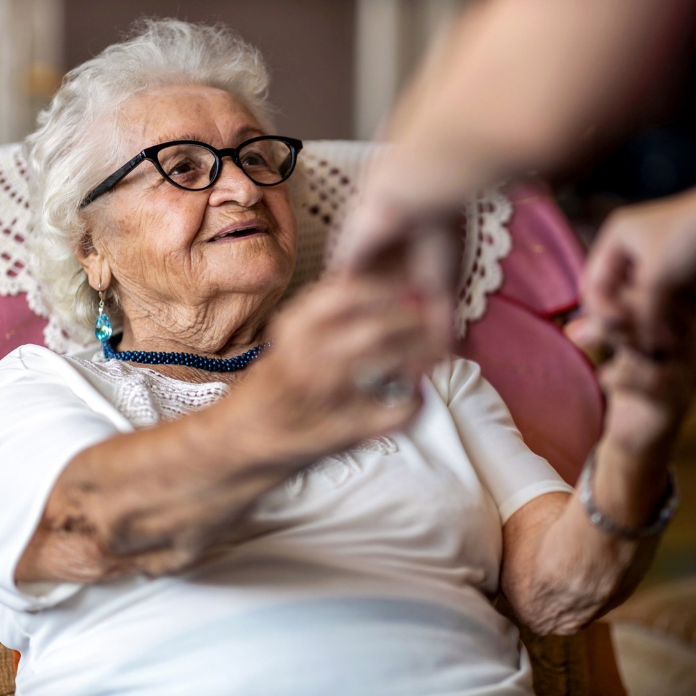 Female home carer supporting old woman to stand up from the armchair at care home-1272459744-1