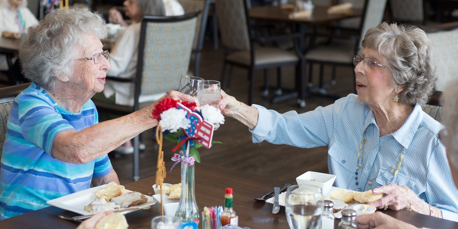 Two seniors toasting with drinks over a meal in the senior living community dining room