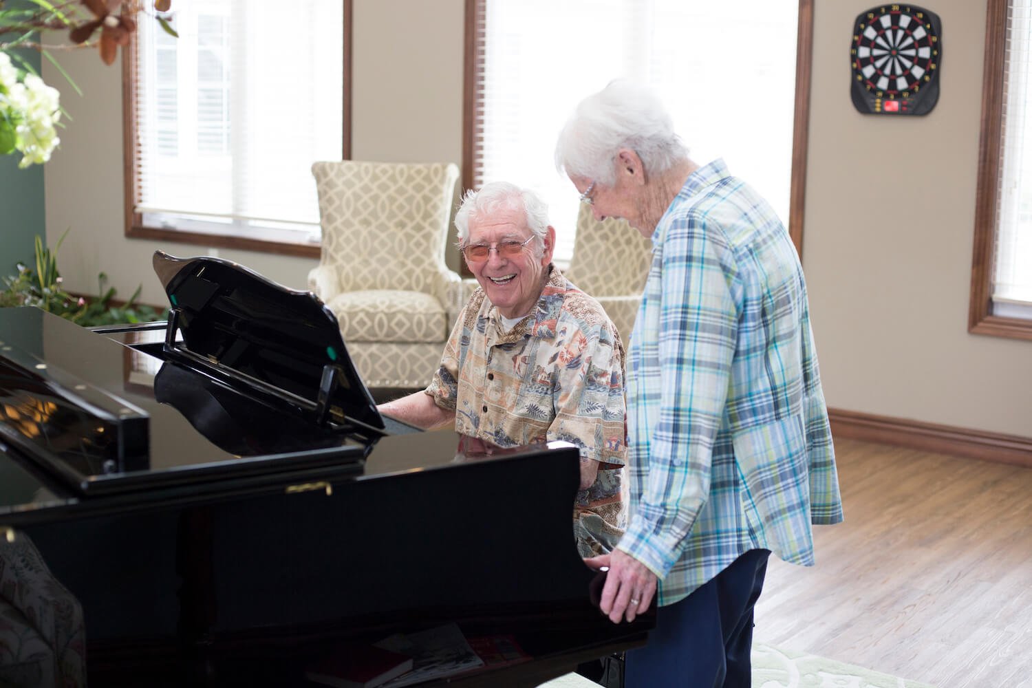 A senior man plays a grand piano while a woman stands beside him