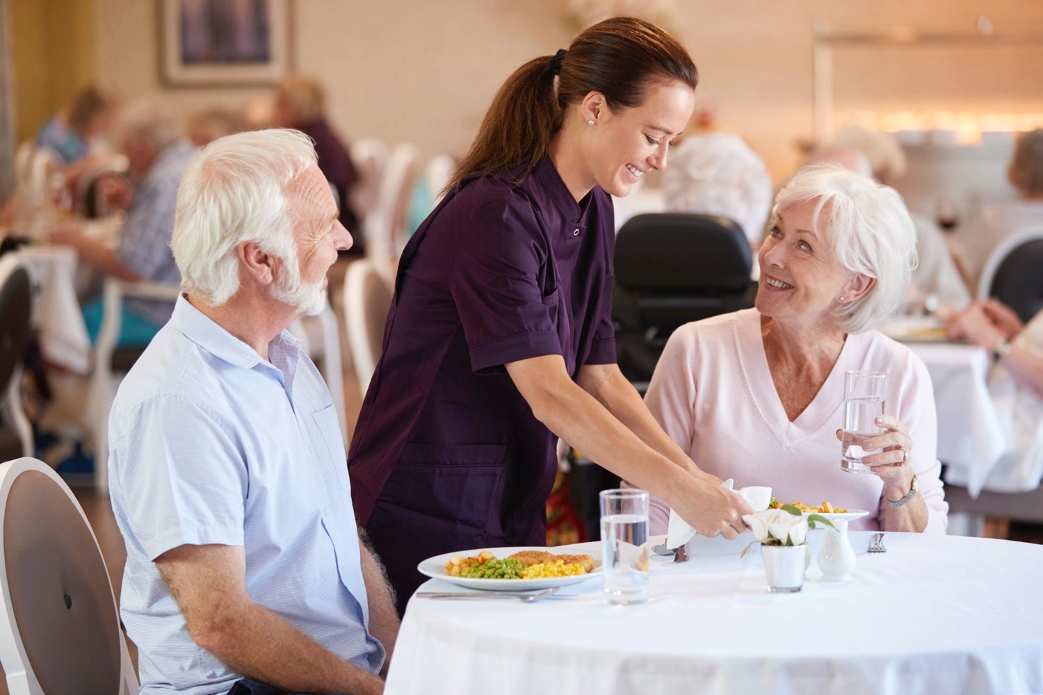 Senior Couple Being Served With Meal By Carer In Dining Room Of Retirement Home-1047536904