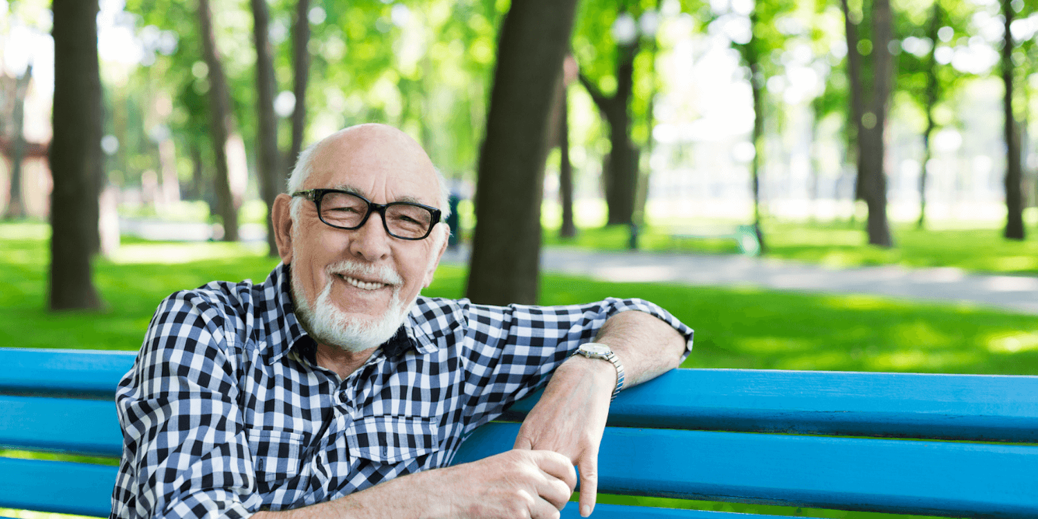 A senior man sits on a blue bench in a sunny park