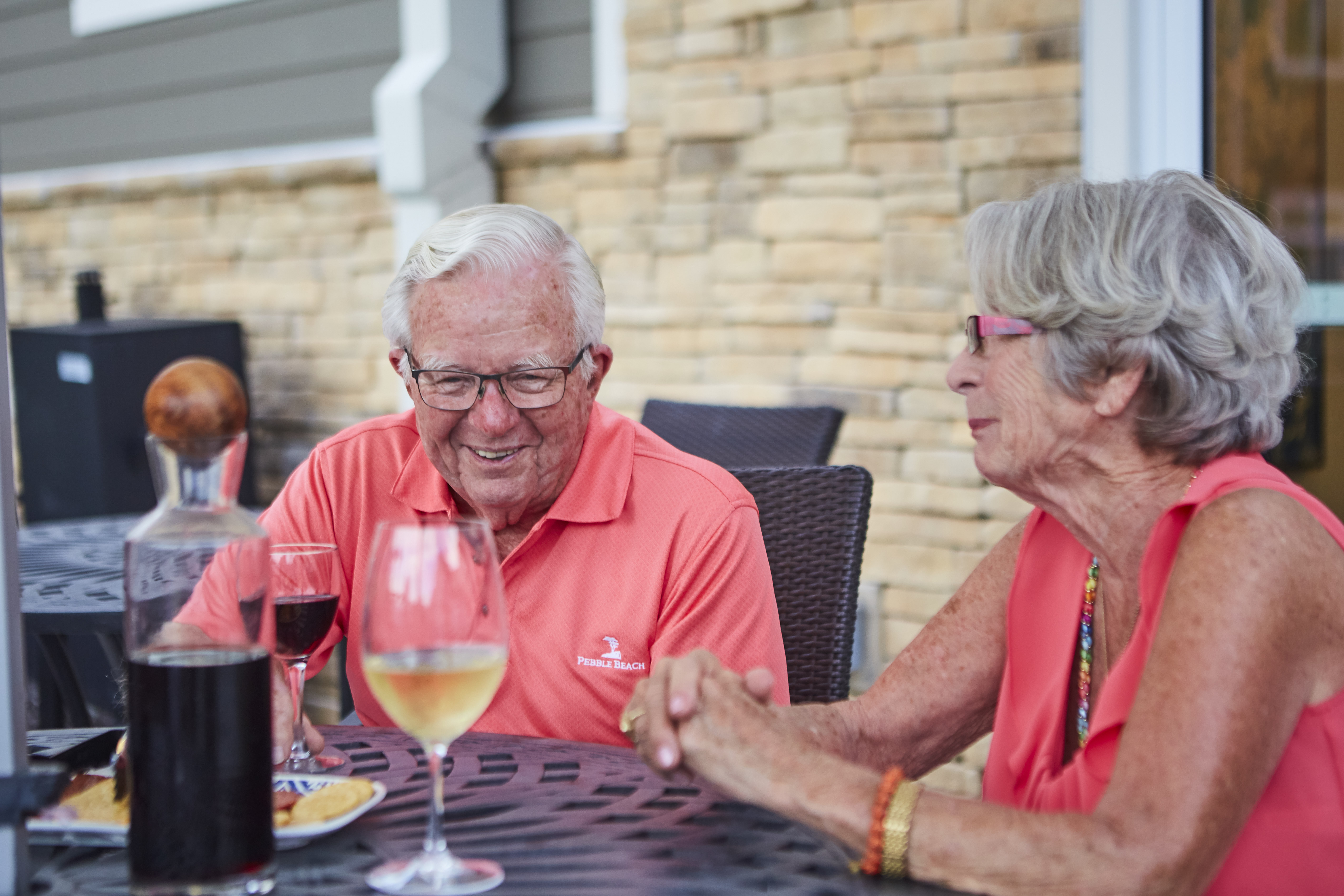 Two seniors drinking wine and eating at an outdoor dining table at a senior living community