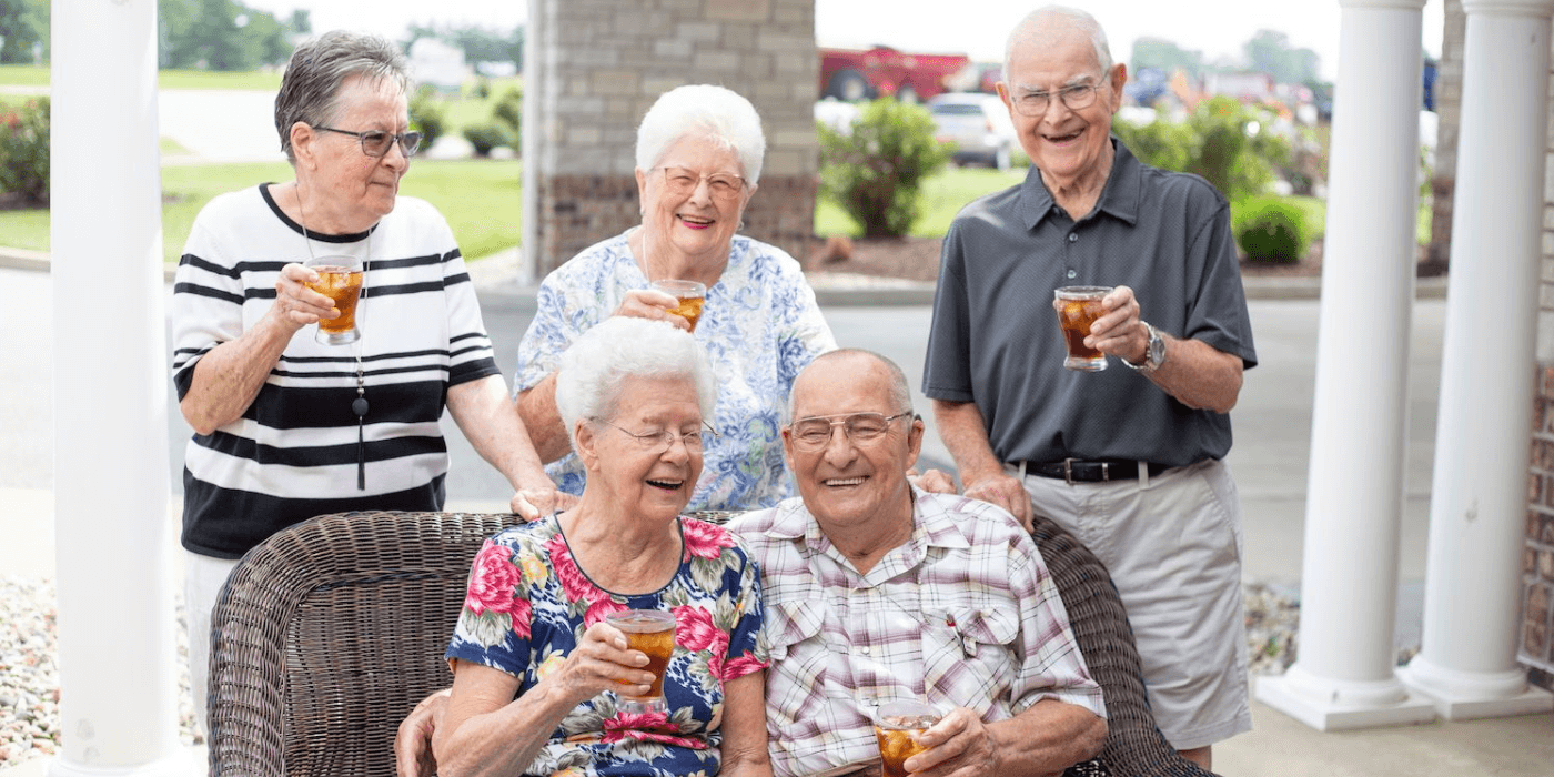 A group of laughing seniors with beverages on a patio in a senior living community.