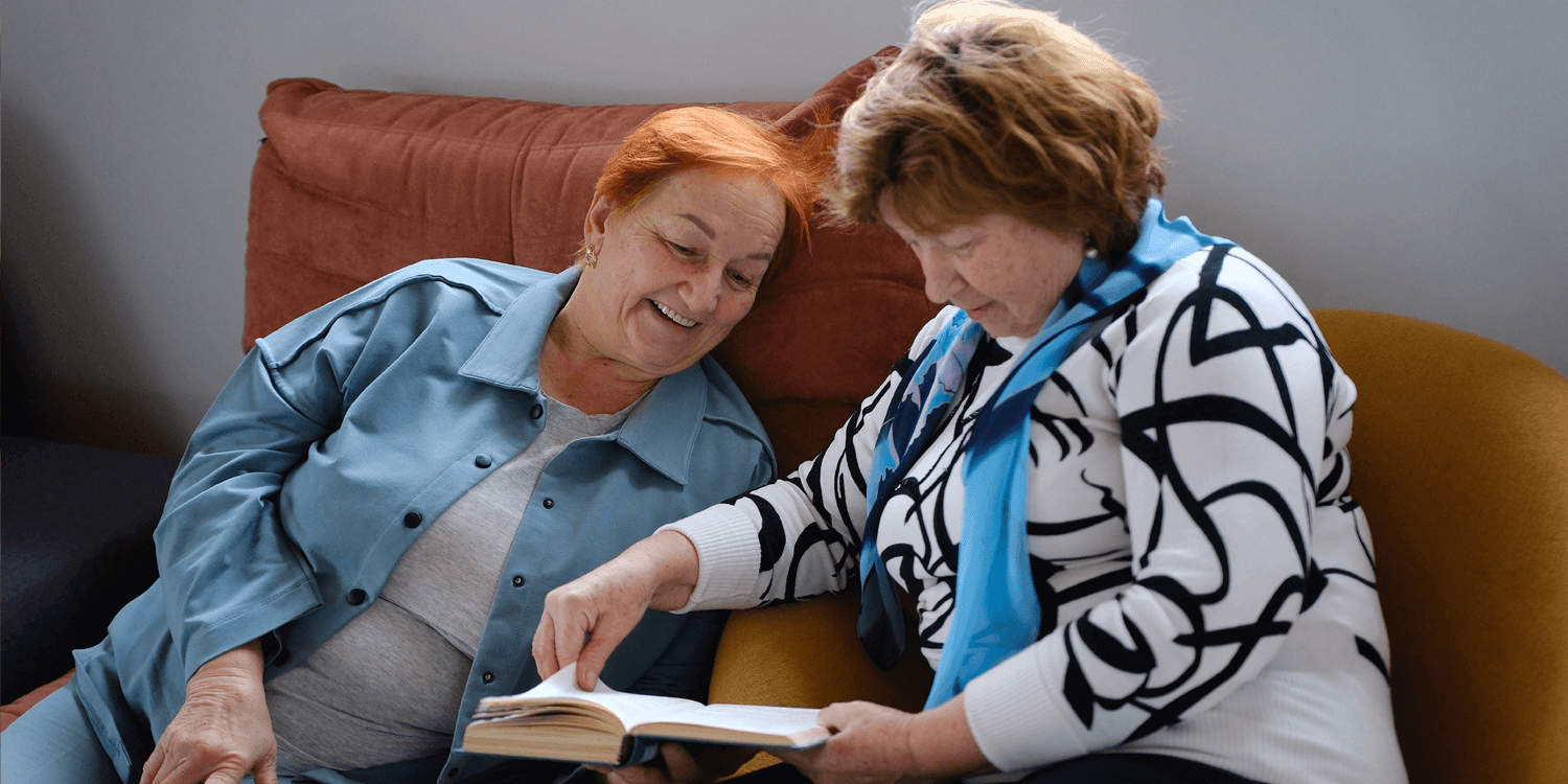 Two assisted living residents sitting on a couch looking at a book