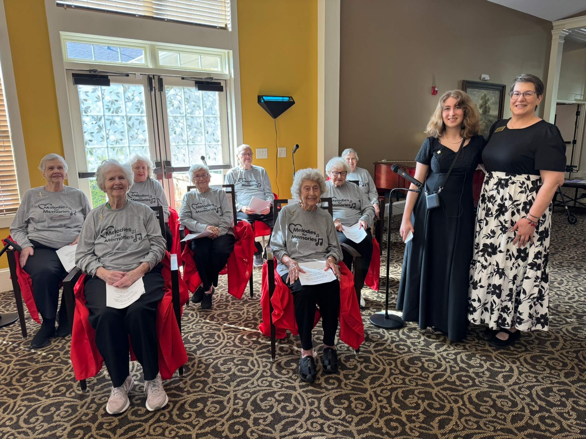 A choir club at a senior living community gathers and poses for a photo. The choir members all wear grey shirts that read "Melodies for Memories."