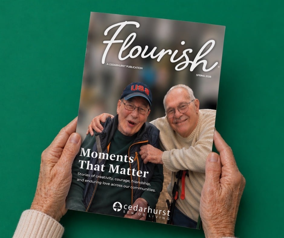 A photo of a senior woman's hands holding an issue of a magazine. The cover features two older adult men with their arms around each other smiling. The title reads 'Flourish Magazine: A Cedarhurst Publication'