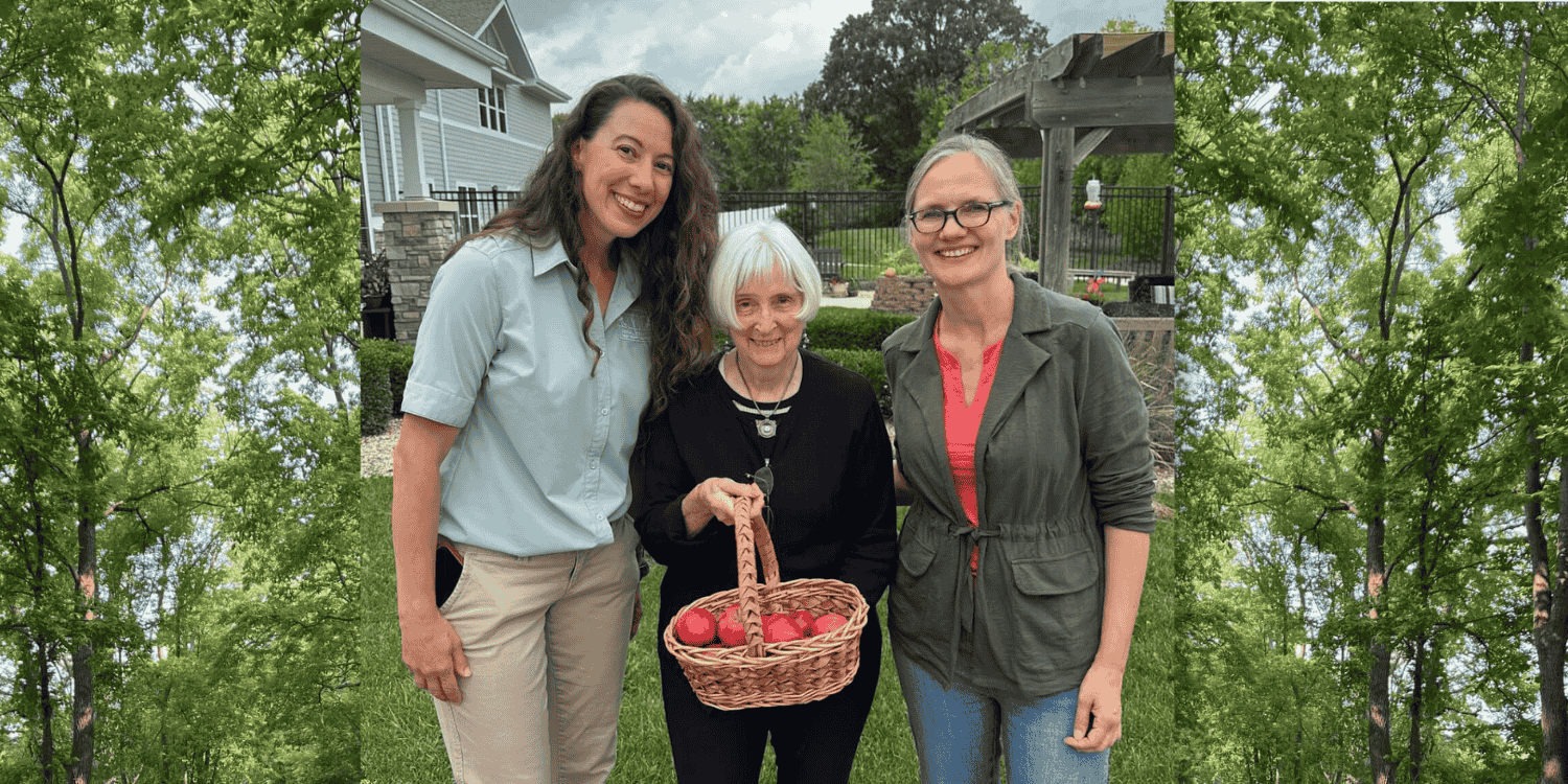 A senior woman holds a wicker basket of apples and smiles, posing for a photo with two women: the Life Enrichment Coordinator of her senior living community and the Head of Education at her community's local Forest Preserve District.