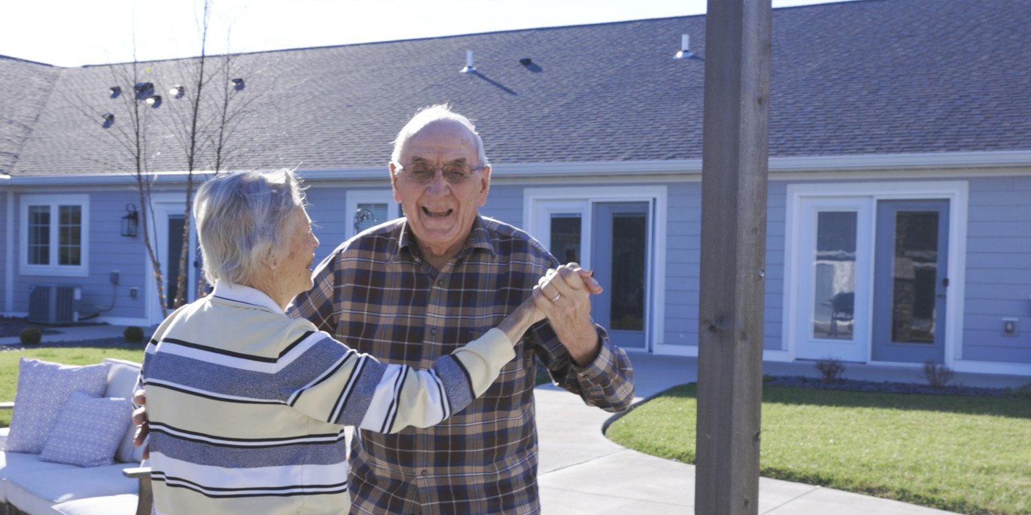 Two seniors happily dancing outdoors and smiling at the camera at an independent living community
