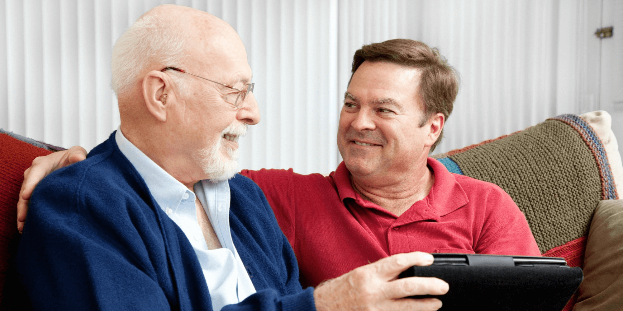 Two people sitting on a sofa and smiling at each other as they read a tablet together.