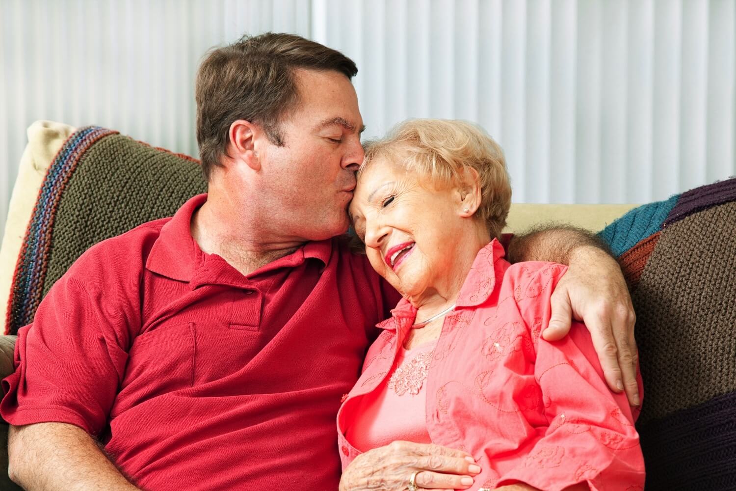 A man kissing his elderly mother's forehead as they sit together on a couch