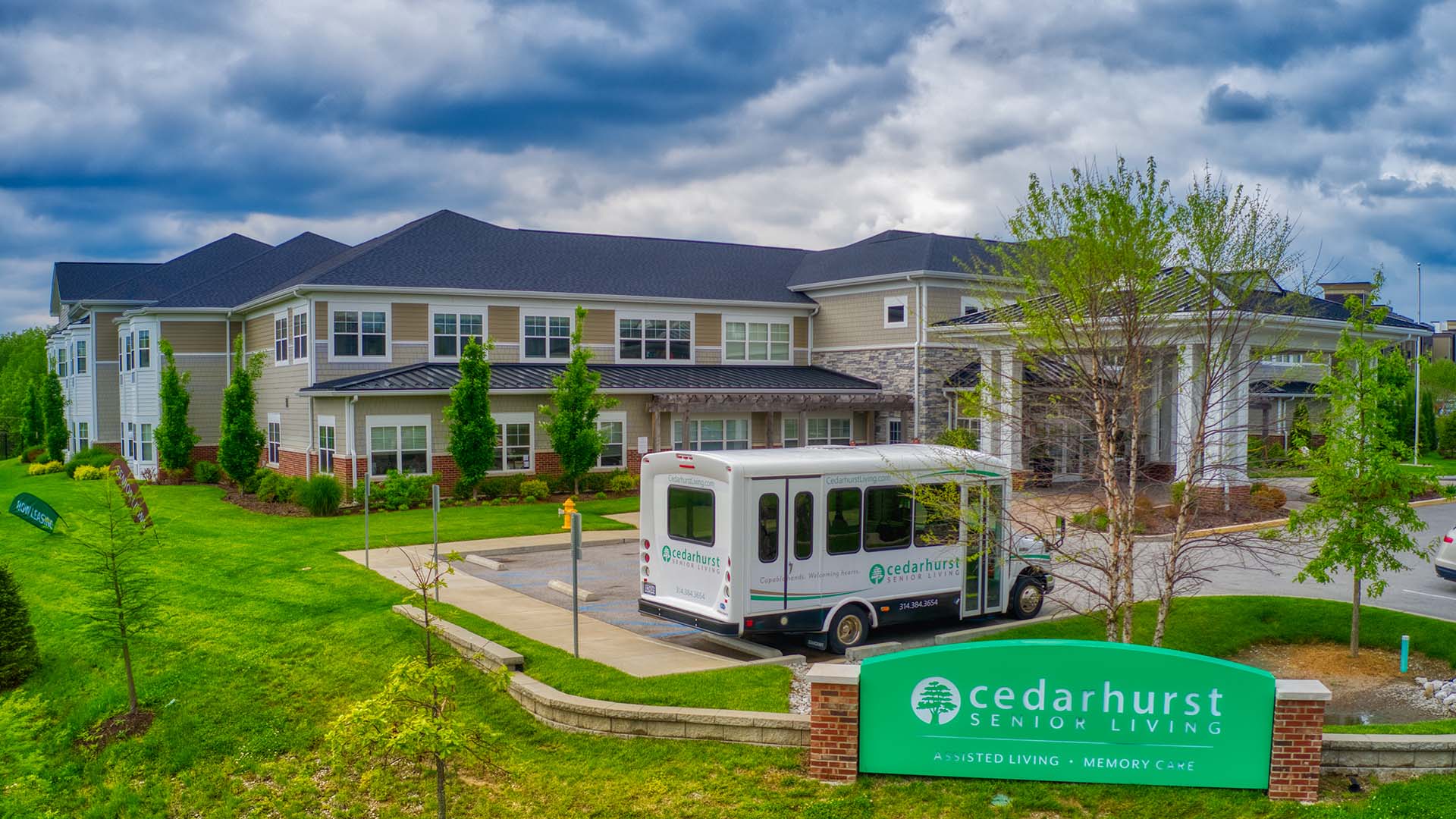 Two-story senior living facility with a Cedarhurst shuttle bus parked outside. Lush greenery surrounds the building, under a slightly cloudy sky