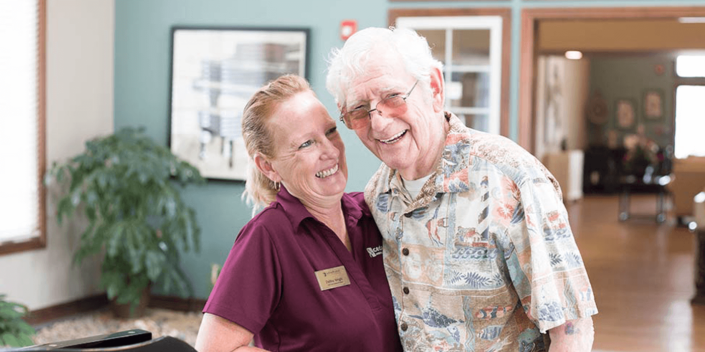 An employee at Cedarhurst, a senior living community, hugs and laughs with an older senior man.