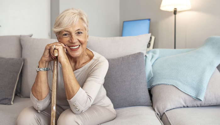 Radiant senior woman sitting on a couch and smiling with cane